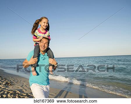 Stock Photo - Hispanic father carrying daughter on shoulders on beach. Fotosearch