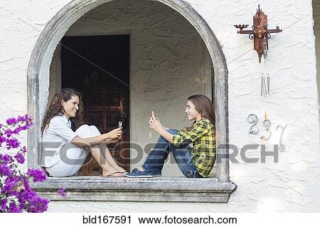 Stock Image - Hispanic mother and daughter using cell phones in window sill. Fotosearch