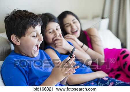 Stock Image - Hispanic siblings using cell phone on sofa. Fotosearch