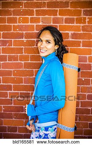 Stock Image - Hispanic woman carrying yoga mat at brick wall. Fotosearch