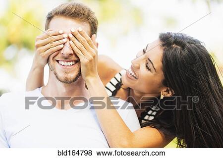 Stock Photo - Hispanic woman covering eyes of boyfriend outdoors. Fotosearch