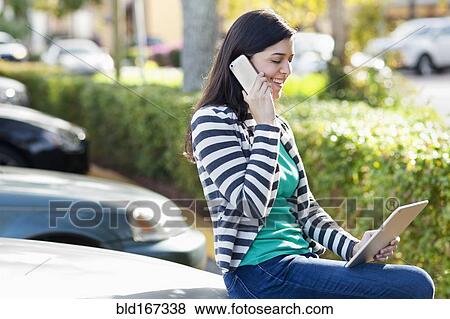 Stock Photo - Hispanic woman using cell phone and digital tablet in parking lot. Fotosearch