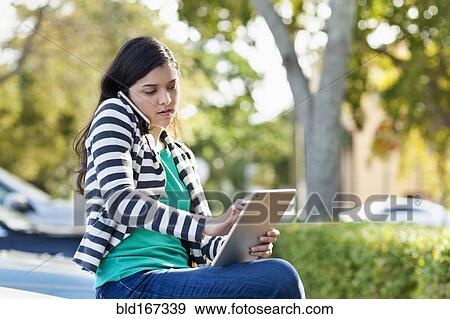 Stock Photo - Hispanic woman using cell phone and digital tablet in parking lot. Fotosearch
