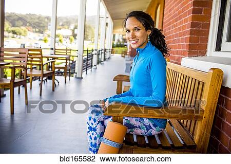 Stock Image - Hispanic woman with yoga mat sitting on bench. Fotosearch