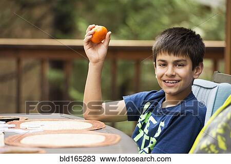 Stock Image - Mixed race boy eating fruit at table on porch. Fotosearch