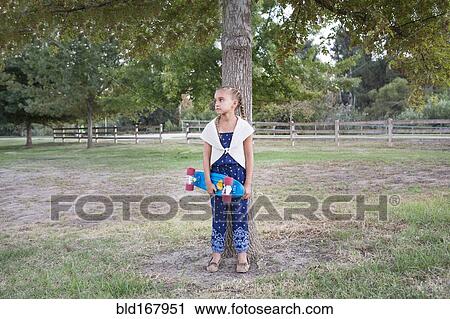 Stock Image - Mixed race girl holding skateboard at tree. Fotosearch