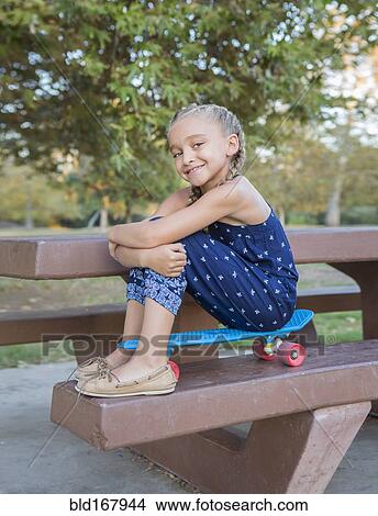 Picture - Mixed race girl sitting on skateboard on picnic table. Fotosearch