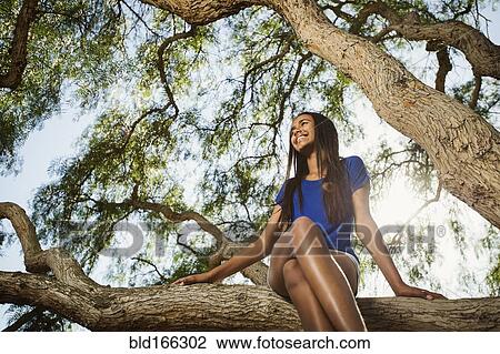 Stock Image - Mixed race girl sitting on tree branch. Fotosearch