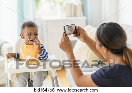 Stock Photograph - Mixed race mother photographing messy baby boy in high chair. Fotosearch