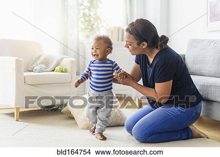 Picture - Mixed race mother playing with baby son in living room. Fotosearch