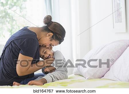 Stock Photo - Mixed race mother tickling baby son on bed. Fotosearch