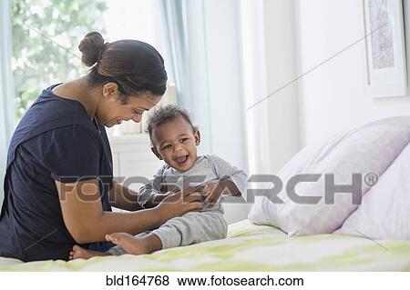 Stock Photo - Mixed race mother tickling baby son on bed. Fotosearch