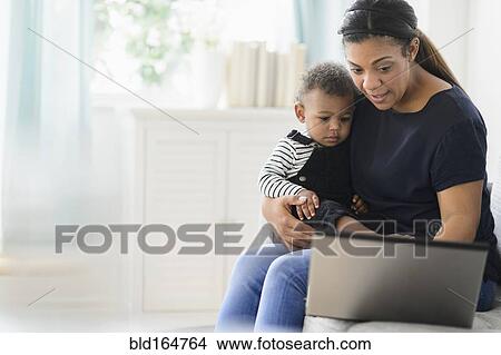 Mixed race mother using laptop with baby son in living room View Large Photo Image Picture - Mixed race mother using laptop with baby son in living room. Fotosearch