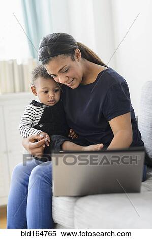 Stock Photography - Mixed race mother using laptop with baby son in living room. Fotosearch