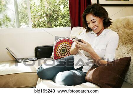 Stock Image - Mixed race woman using cell phone on sofa. Fotosearch