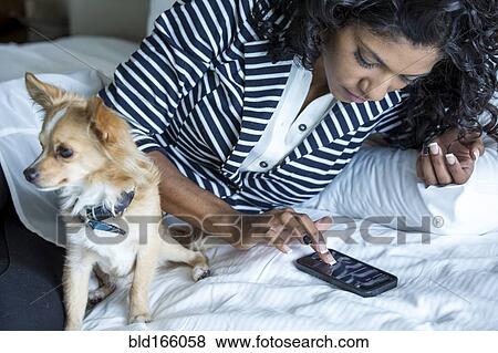 Stock Photo - Mixed race woman using cell phone with dog on bed. Fotosearch