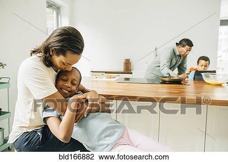 Stock Image - Mother hugging daughter in kitchen. Fotosearch
