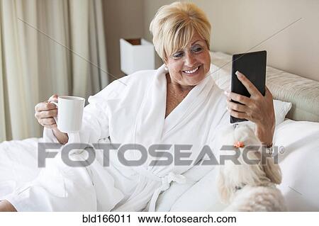 Stock Image - Older Caucasian woman and dog using digital tablet on bed. Fotosearch