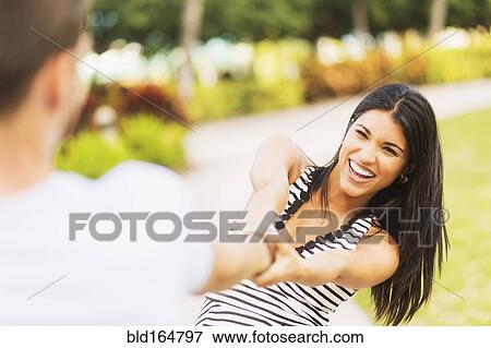 Stock Photo - Smiling Hispanic woman pulling boyfriend in park. Fotosearch