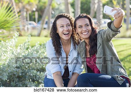 Stock Photo - Smiling women taking selfie in park. Fotosearch
