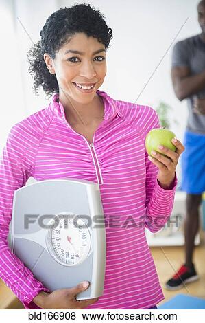 Stock Photo - Woman holding apple and scale in gym. Fotosearch