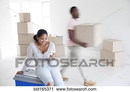 Stock Image - Woman relaxing with man carrying boxes in new home. Fotosearch