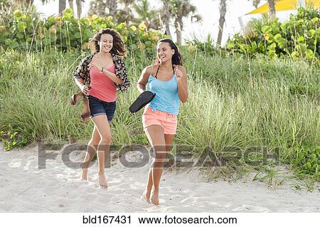 Stock Image - Women carrying shoes running on beach. Fotosearch