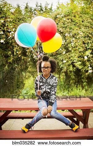 Stock Image - Black girl holding bunch of balloons on picnic table. Fotosearch