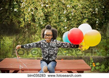 Stock Image - Black girl holding bunch of balloons on picnic table. Fotosearch