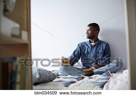 Stock Photo - Black man drinking tea and reading on bed. Fotosearch
