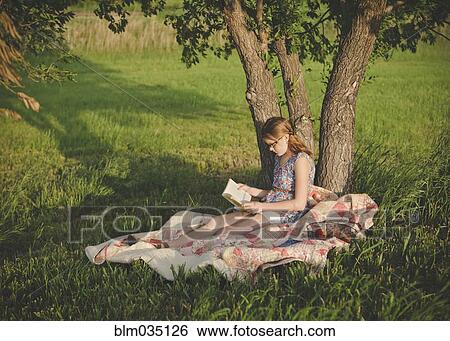 Stock Photograph - Caucasian girl reading book under tree in field. Fotosearch