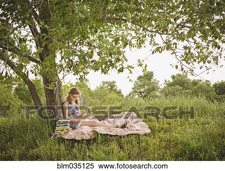 Stock Photography - Caucasian girl reading book under tree in field. Fotosearch