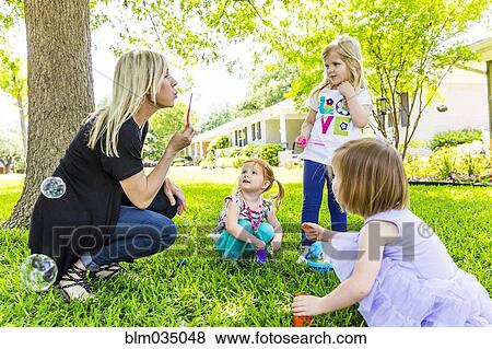 Stock Photo - Caucasian mother and daughters blowing bubbles in backyard. Fotosearch