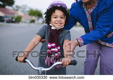 Stock Image - Grandmother teaching granddaughter to ride bicycle on suburban street. Fotosearch
