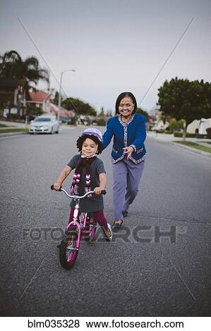 Stock Photo - Grandmother teaching granddaughter to ride bicycle on suburban street. Fotosearch