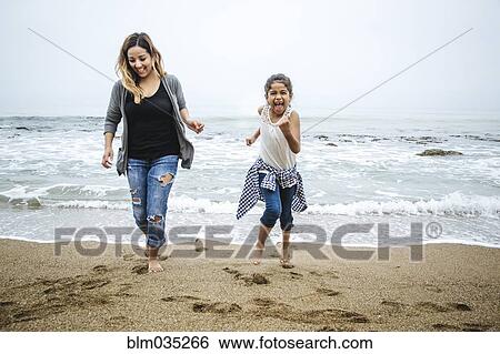 Stock Photograph - Hispanic mother and daughter walking barefoot on beach. Fotosearch
