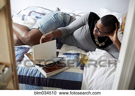 Stock Image - Mixed race woman drinking tea and reading on bed. Fotosearch