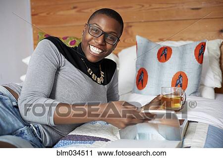 Stock Image - Mixed race woman drinking tea and reading on bed. Fotosearch