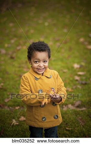 Stock Image - Smiling girl wearing yellow coat holding autumn leaves. Fotosearch