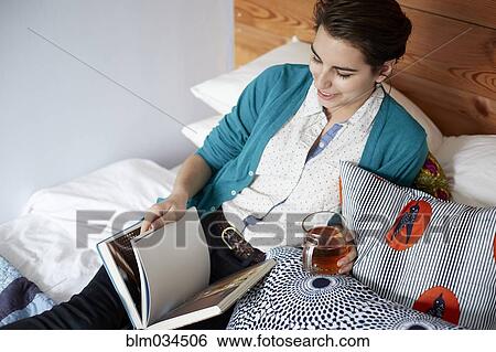 Stock Photograph - Woman drinking tea and reading in bed. Fotosearch