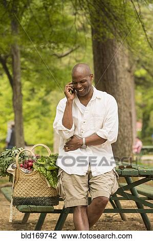 Stock Image - African American man talking on cell phone on picnic table. Fotosearch