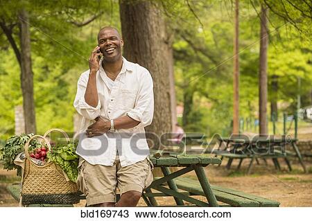 Stock Image - African American man talking on cell phone on picnic table. Fotosearch