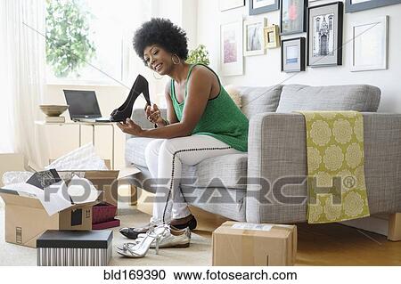 Stock Image - African American woman opening packages of shoes on sofa. Fotosearch
