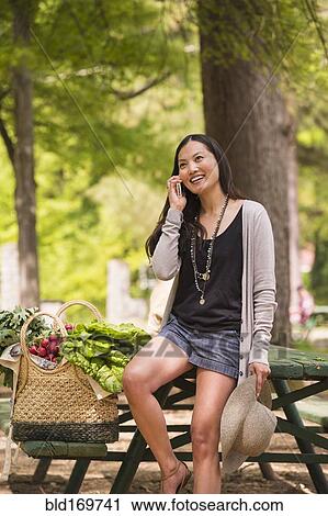 Stock Image - Asian woman talking on cell phone on picnic table. Fotosearch