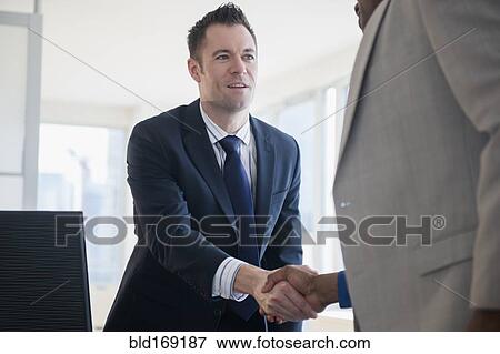 Stock Photo - Businessmen shaking hands at desk in office. Fotosearch