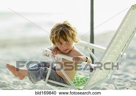 Stock Image - Caucasian baby boy sitting in lawn chair on beach. Fotosearch