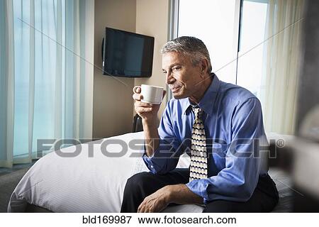 Stock Photo - Caucasian businessman drinking coffee on hotel bed. Fotosearch
