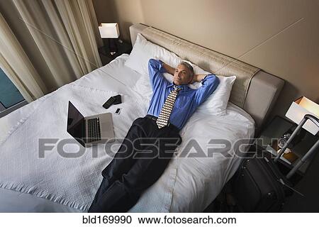 Stock Image - Caucasian businessman laying with laptop on hotel bed. Fotosearch