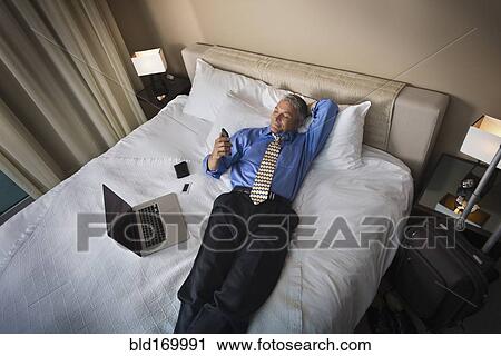 Stock Image - Caucasian businessman using cell phone on hotel bed. Fotosearch