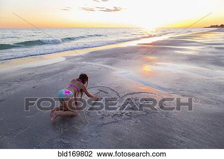 Stock Image - Caucasian girl drawing in sand on beach. Fotosearch